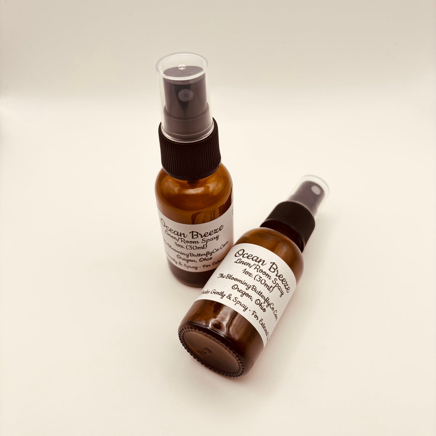 To amber colored spray, bottles of ocean breeze, linen and room spray with white labels handcrafted by the blooming butterfly company