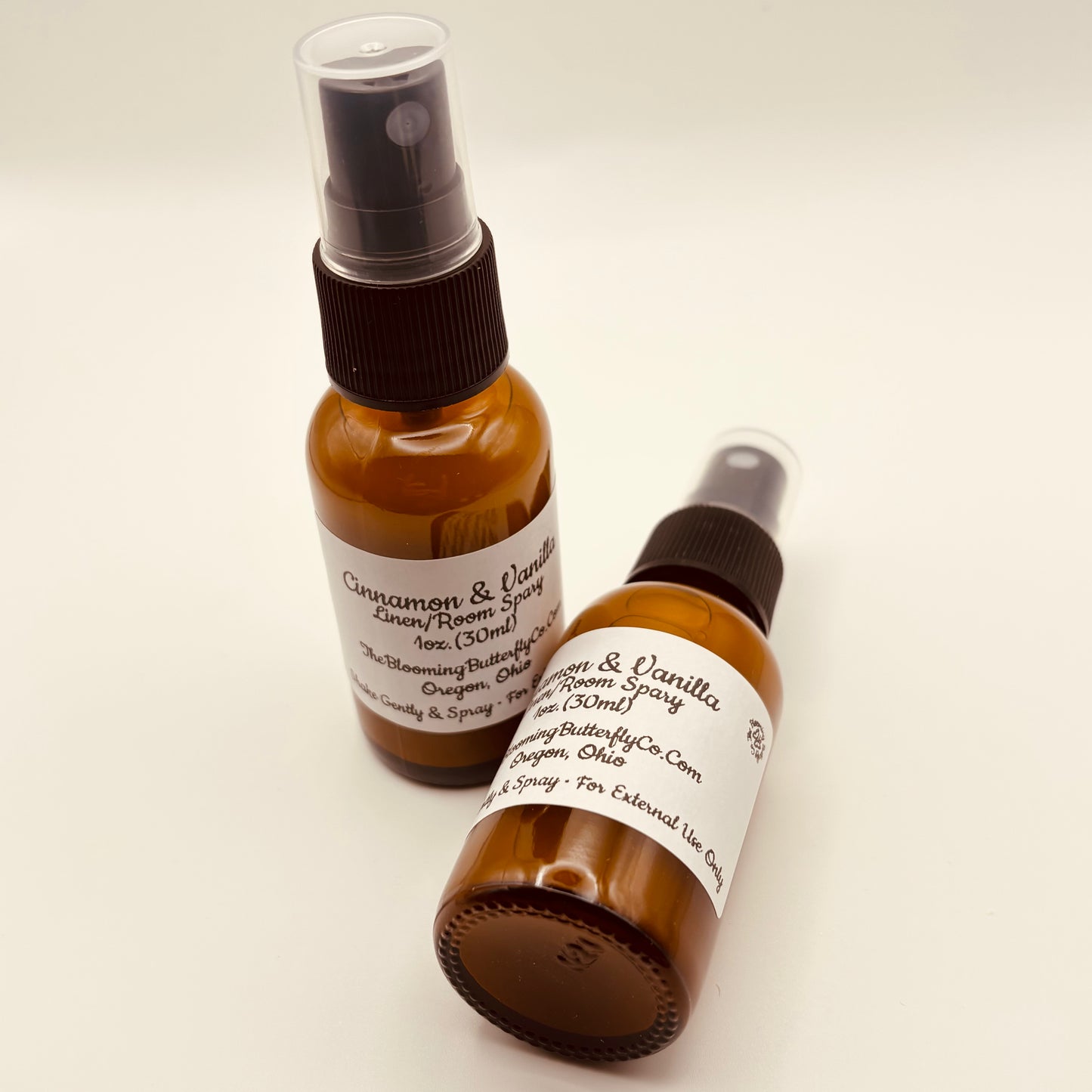 Two Amber colored spray, bottles of cinnamon and vanilla linen room spray with white labels handcrafted by the blooming butterfly company