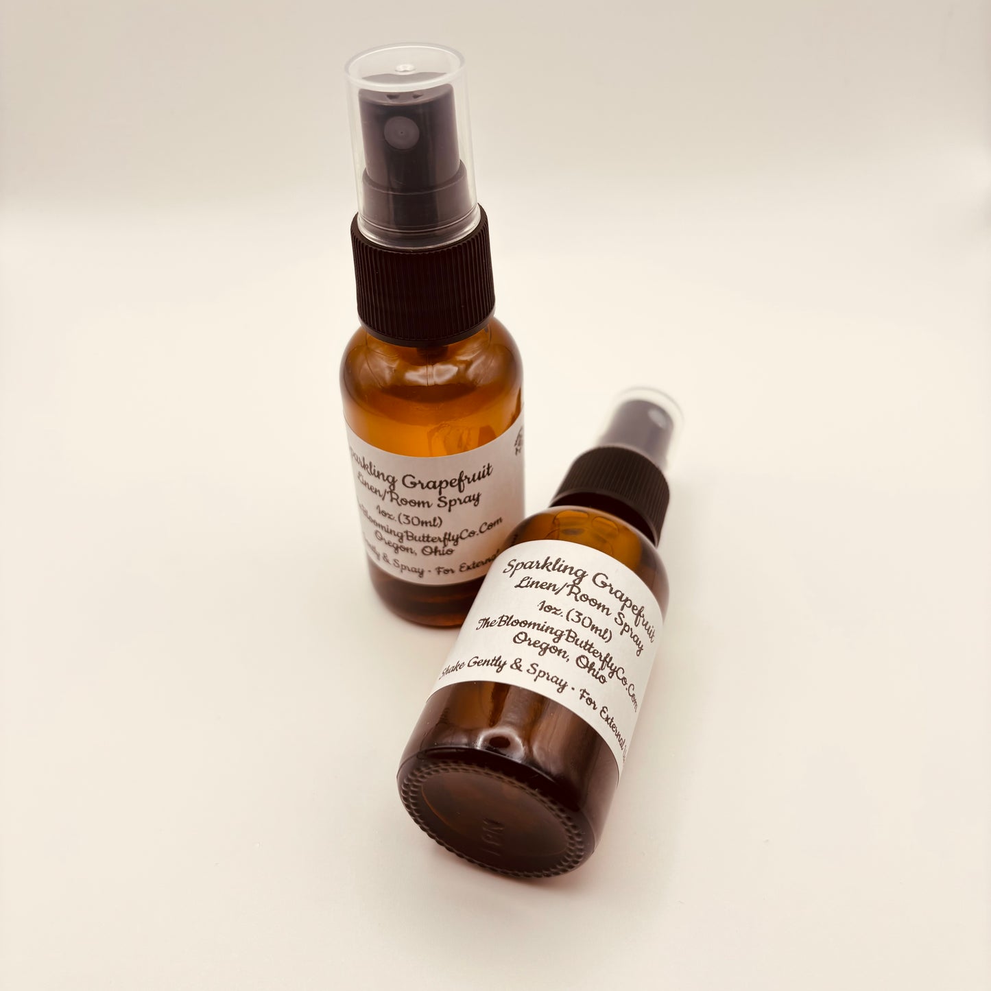Two amber colored spray bottles of sparkling grapefruit linen/room spray with white labels handcrafted by the blooming butterfly company