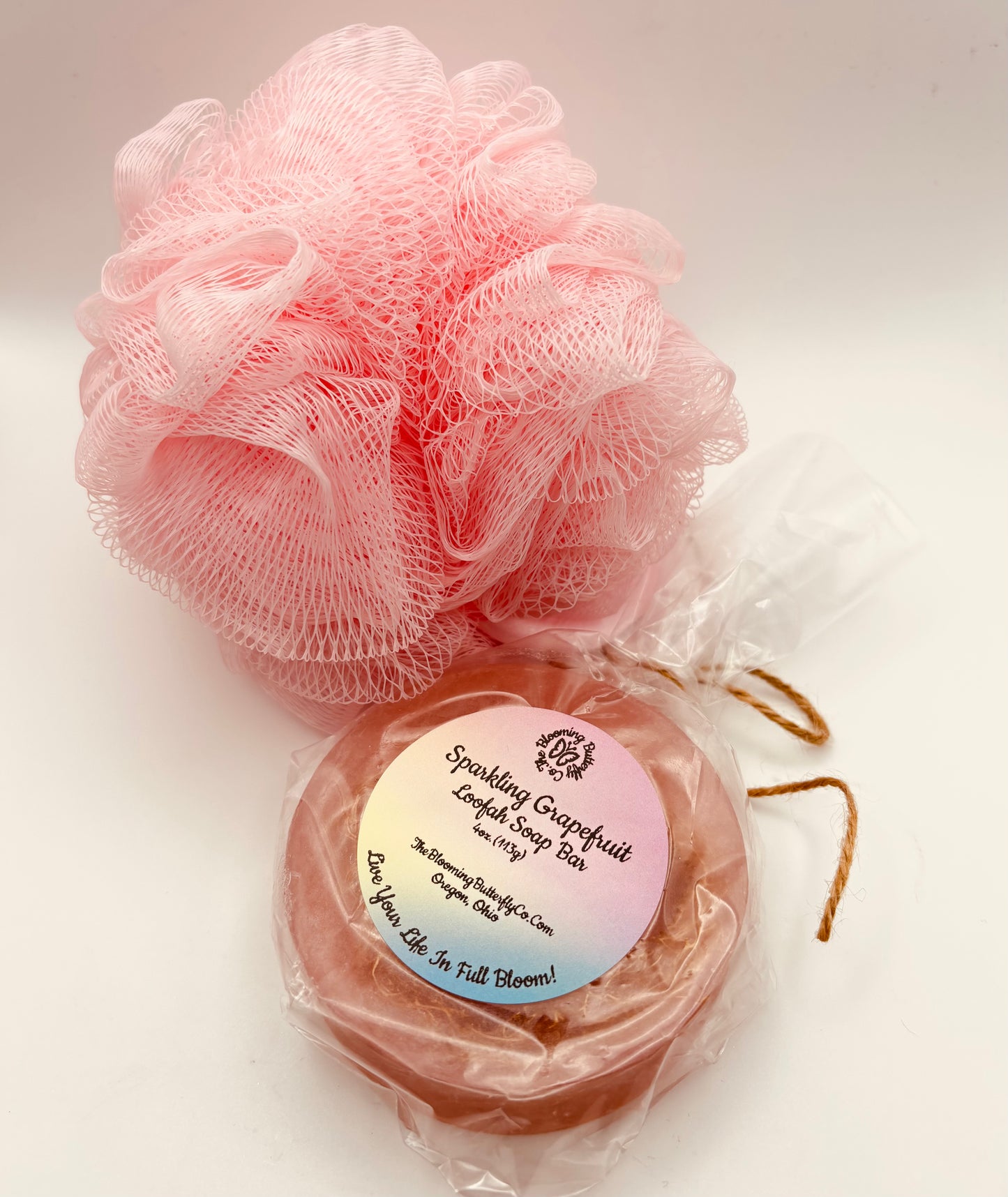 a loofah bath sponge and a round pink sparkling grapefruit loofah soap bar wrapped in cellophane with brown twine handcrafted by the blooming butterfly company