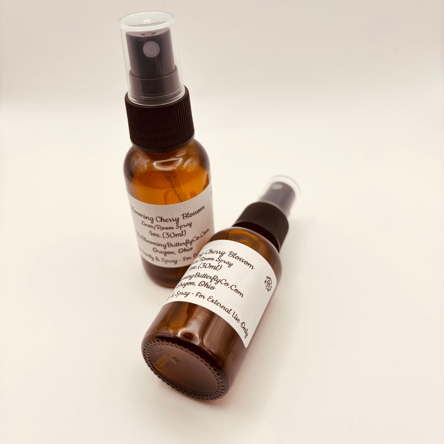 Two Amber colored spray bottles of blooming cherry blossom linen room spray with white labels created by the blooming butterfly company
