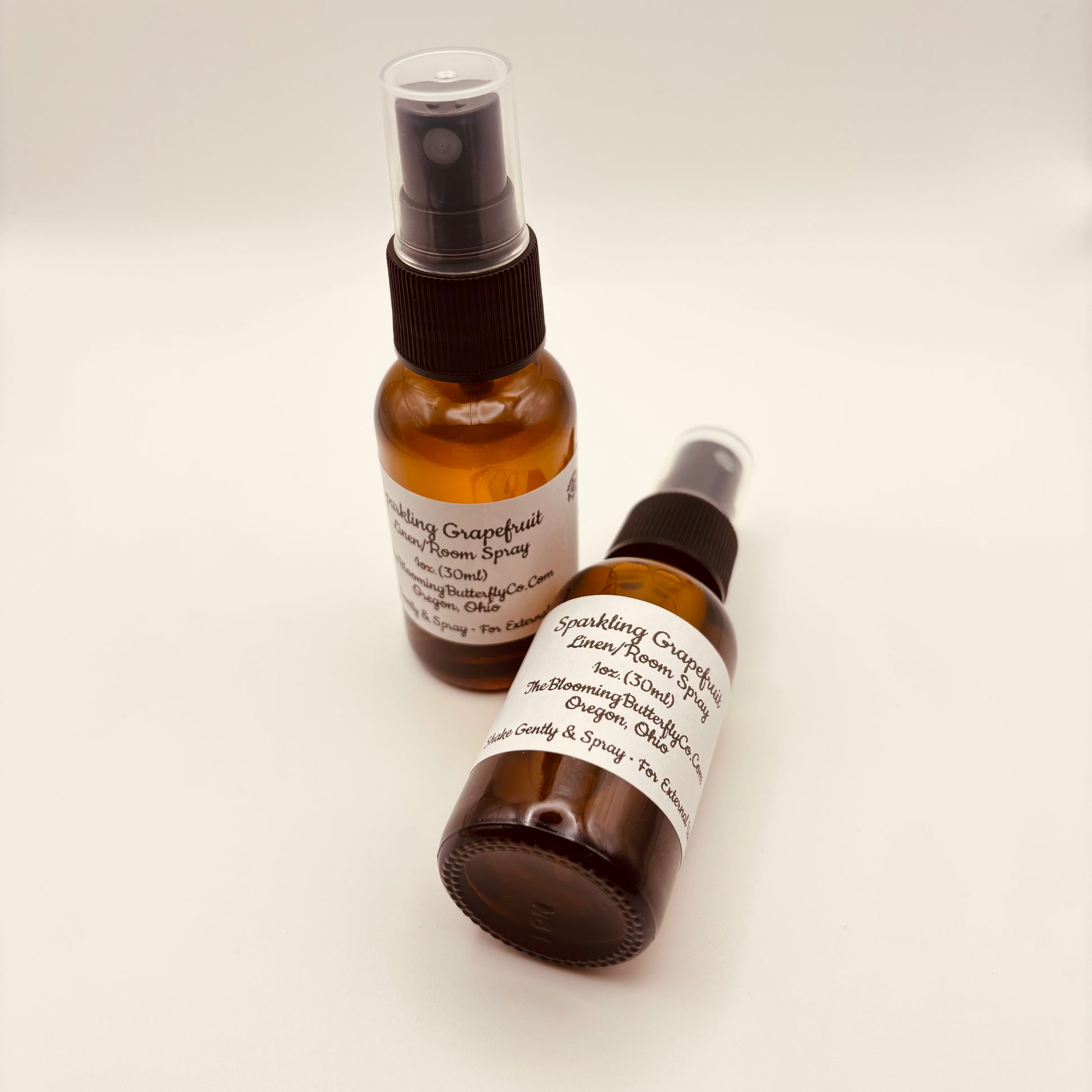 Two amber colored spray bottles of sparkling grapefruit linen/room spray with white labels handcrafted by the blooming butterfly company