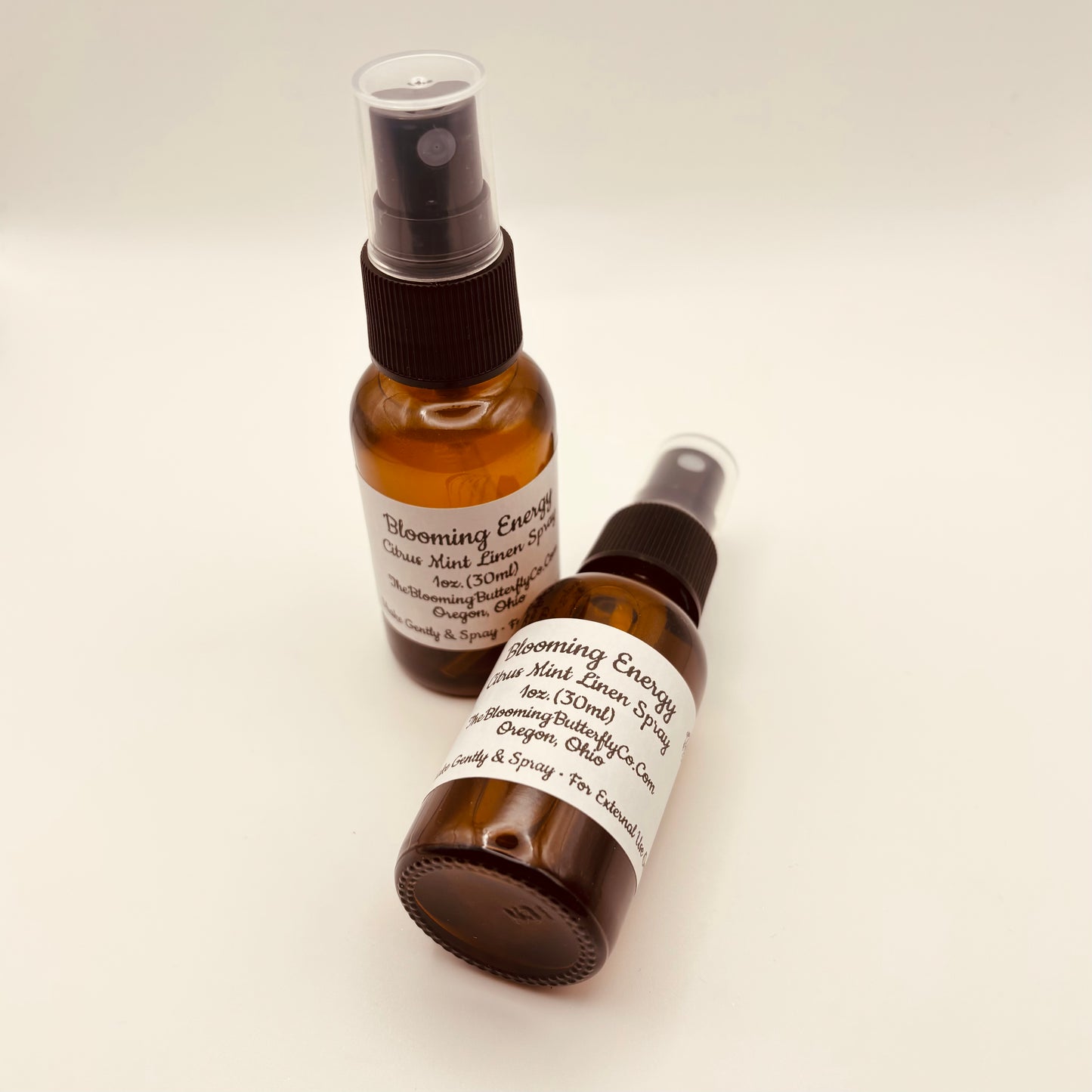 Two Amber colored spray bottles of blooming energy, citrus, mint linen, and room spray with white labels handcrafted by the blooming butterfly company