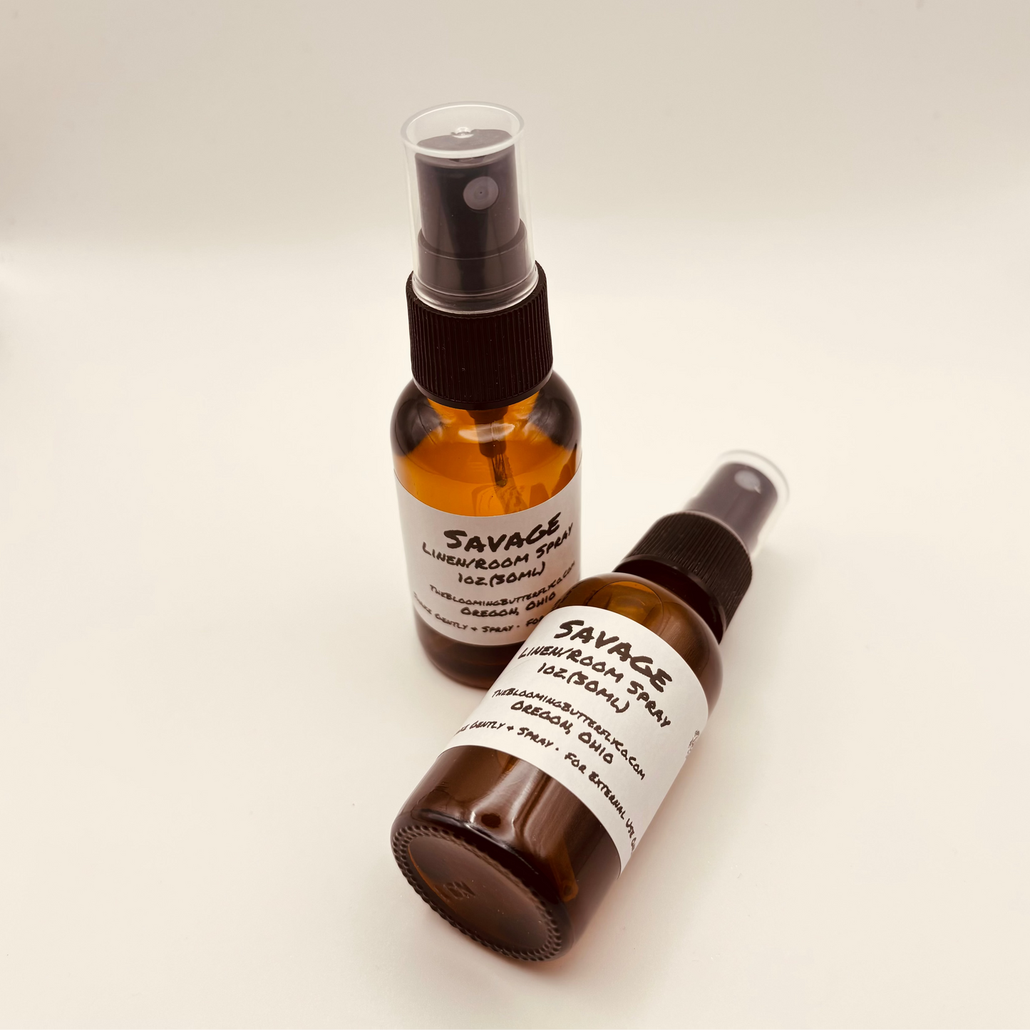 Two Amber colored spray bottles Of Savage type fragrance linen and room spray with white labels and handcrafted by the blooming butterfly Company