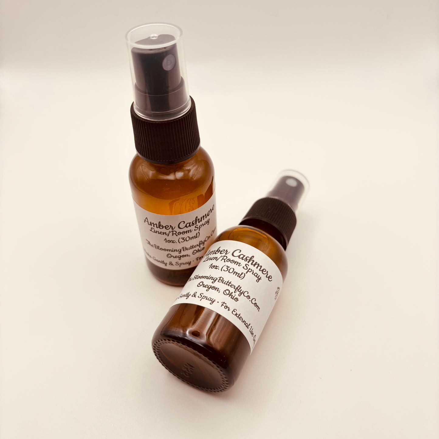 Two Amber colored spray bottles of amber, cashmere linen room spray with white label, handcrafted by the blooming butterfly company
