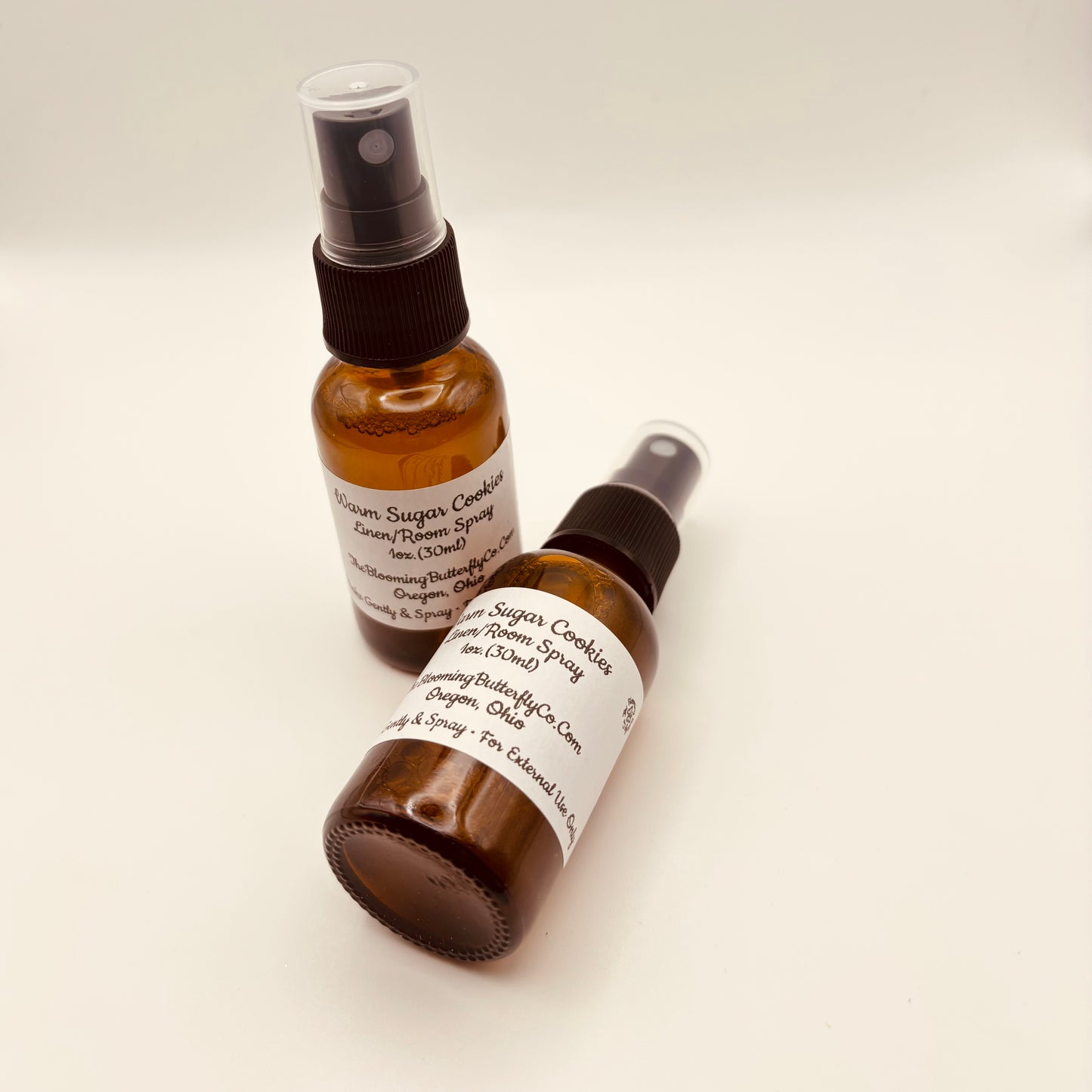 Two Amber colored Spray bottles Of warm sugar, cookie linen room spray with labels handcrafted by the blooming butterfly company