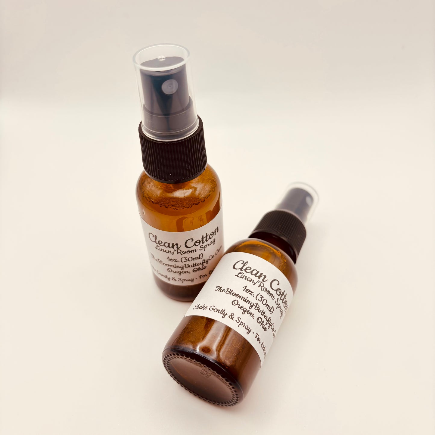 Two Amber colored spray bottles of clean cotton linen/room spray with white labels handmade by the blooming butterfly company