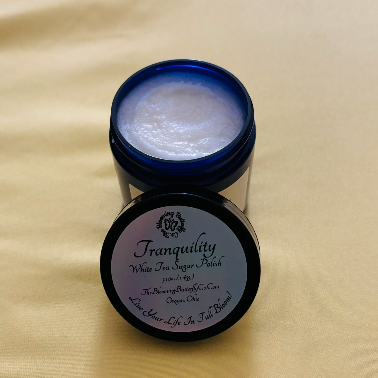 A jar of tranquility, white tea, body polish in a cobalt blue pet jar with a black lid and silver holographic label handcrafted by the blooming butterfly company 5.10 ounces